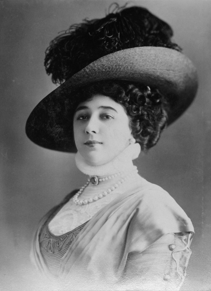 Carolina Otero in a daytime portrait with a wide-brimmed hat and pearls — a far more restrained look than her jewel-covered stage costumes.